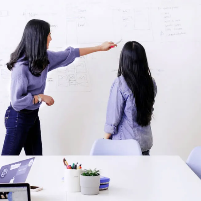 Two women working at a whiteboard in an office space, one pointing to notes on the board while the other observes. The desk in front of them has a laptop and office supplies, indicating a collaborative brainstorming session.