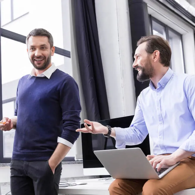 Two men in a professional setting, one holding a laptop, smiling and engaging in conversation. The background features modern office decor with large windows.