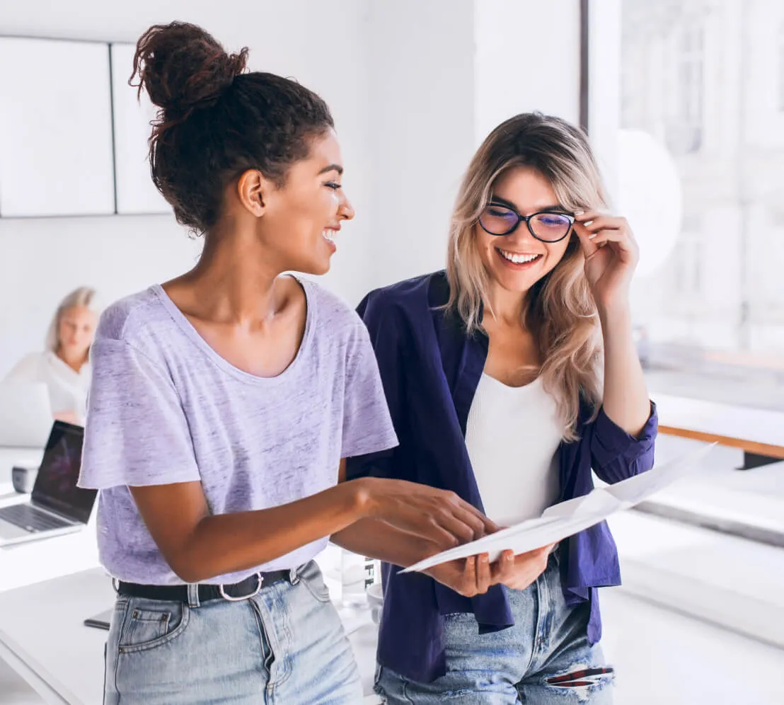 Two women, one holding a document, engage in a conversation in an office. They are smiling, suggesting a light and friendly workplace atmosphere.