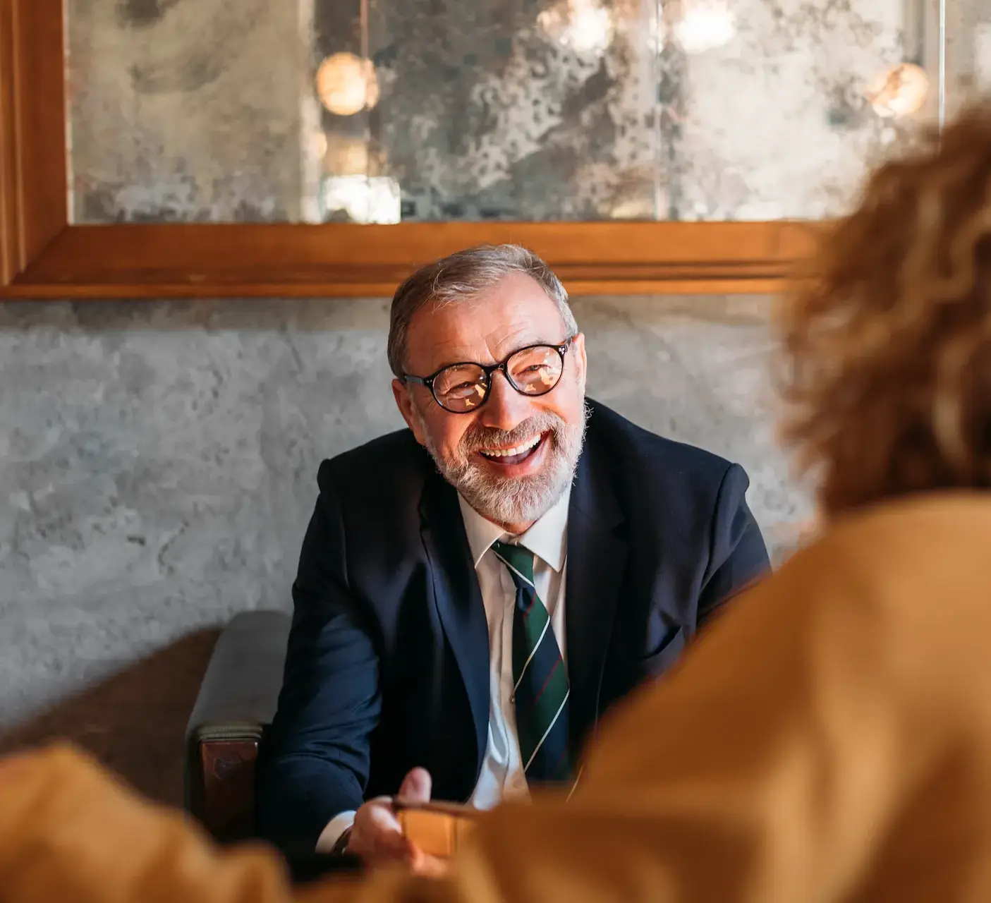 An older man with glasses, a beard, and a joyful smile is engaged in a conversation. He is wearing a dark suit, white shirt, and a green-striped tie. The setting is warm and modern, with a visible wooden panel and textured wall in the background. Part of another person, likely the one he’s conversing with, is blurred in the foreground, enhancing the focus on his cheerful expression.