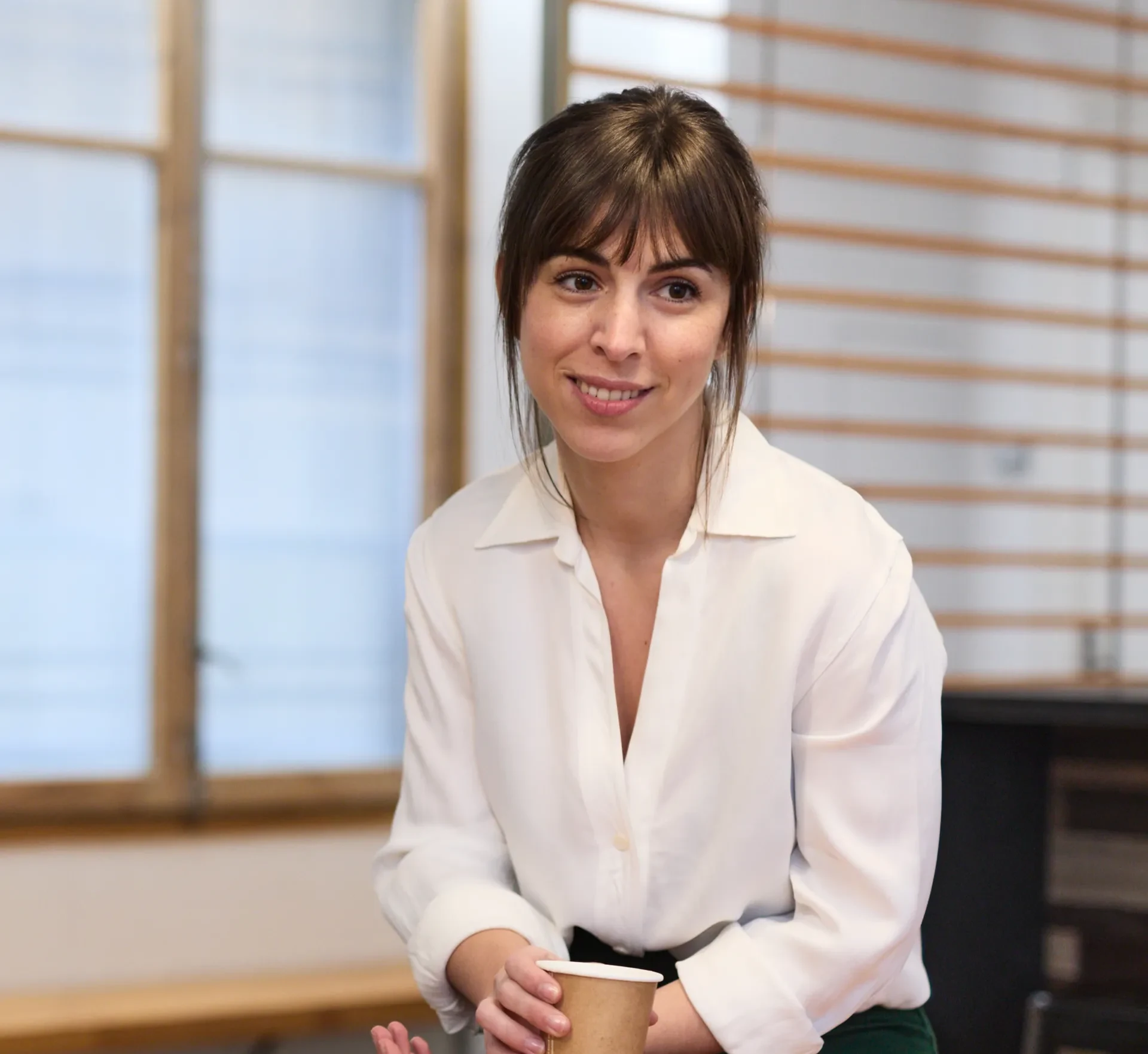 A woman with brown hair tied in a loose updo is sitting and smiling while holding a paper coffee cup. She is wearing a white blouse with an open collar, and her expression is relaxed and friendly. The background includes large windows and a wooden wall, suggesting a modern, minimalistic workspace or meeting area.