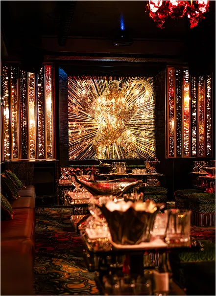 Intérieur d'un bar chic avec une grande œuvre d'art lumineuse dorée représentant une silhouette humaine, des canapés sombres et des tables avec verres et décorations brillantes.