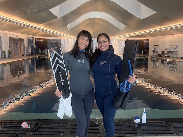 Two women in wetsuits holding diving fins standing by an indoor pool with arched ceiling.