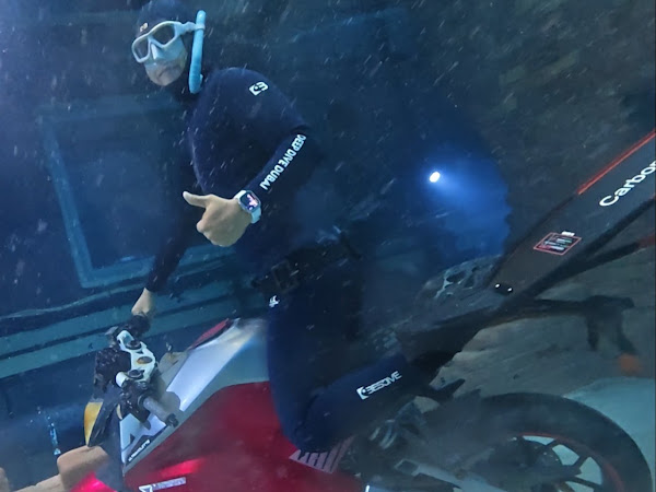 Person underwater wearing wetsuit and snorkel, sitting on a submerged motorcycle, hand showing thumbs up.