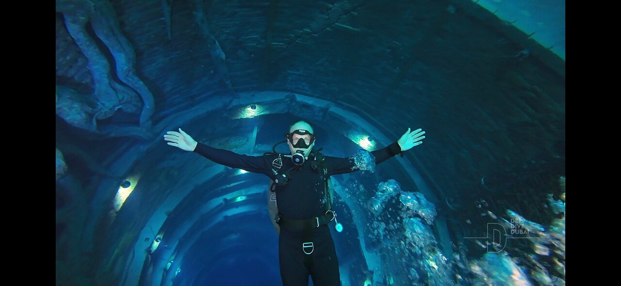 Scuba diver in black wetsuit with arms outstretched inside an underwater tunnel structure.