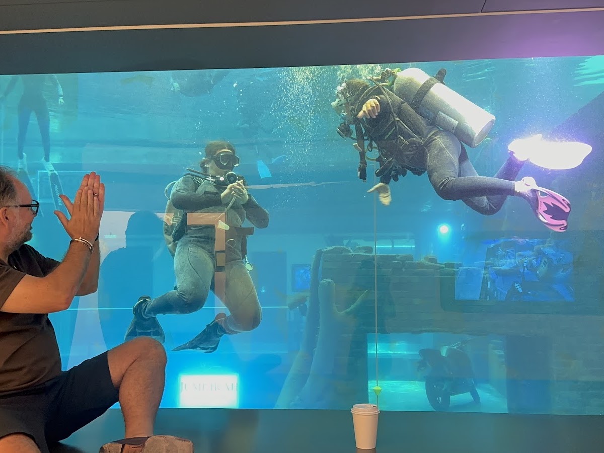 Two scuba divers inside tank with a man outside clapping.
