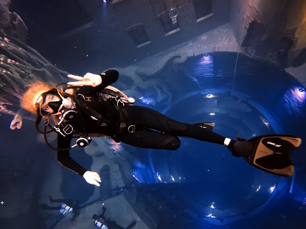 Scuba diver in black wetsuit and gear giving peace sign underwater near sunken structure.