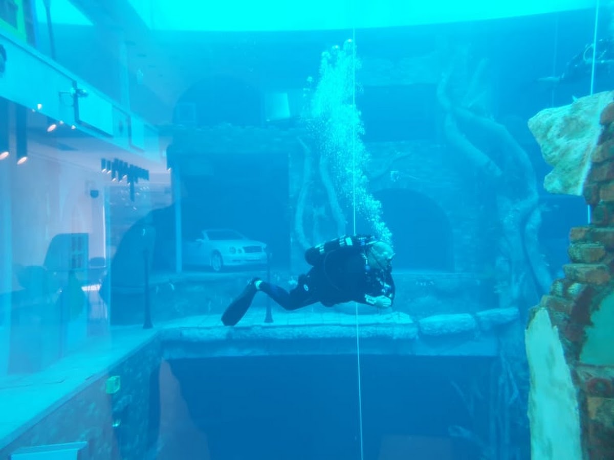 Scuba diver submerged in large aquarium tank with rocks and car inside.