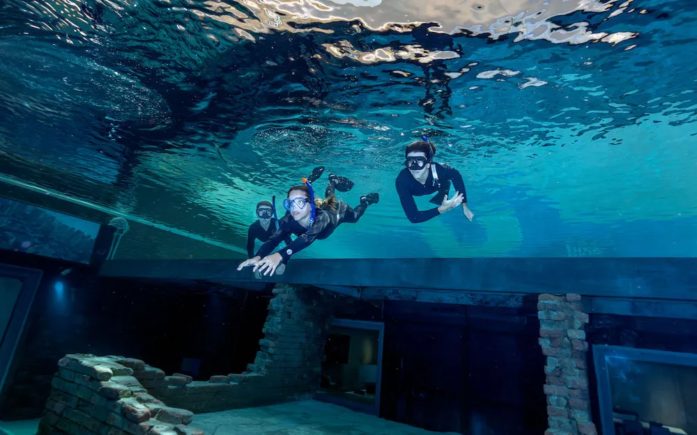Three snorkelers swimming underwater above a submerged brick structure in clear blue water.