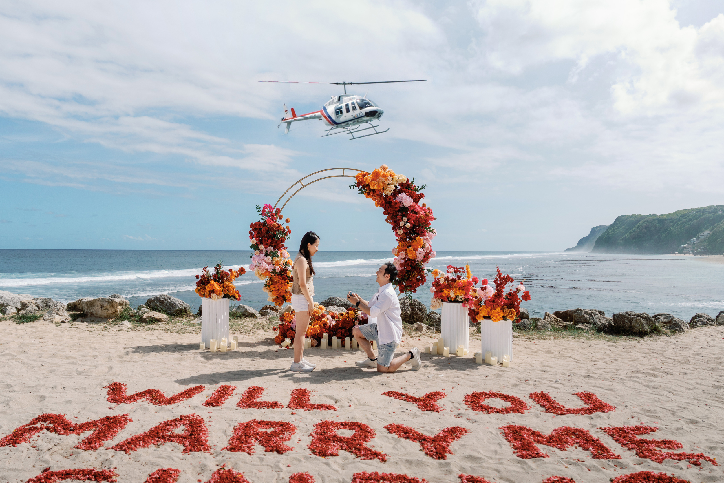 Couple walking towards a cliffside proposal setup with Will You Marry Me written in red rose petals