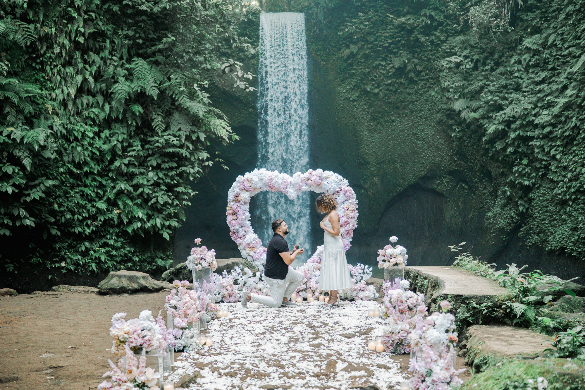 Couple walking towards a cliffside proposal setup with Will You Marry Me written in red rose petals