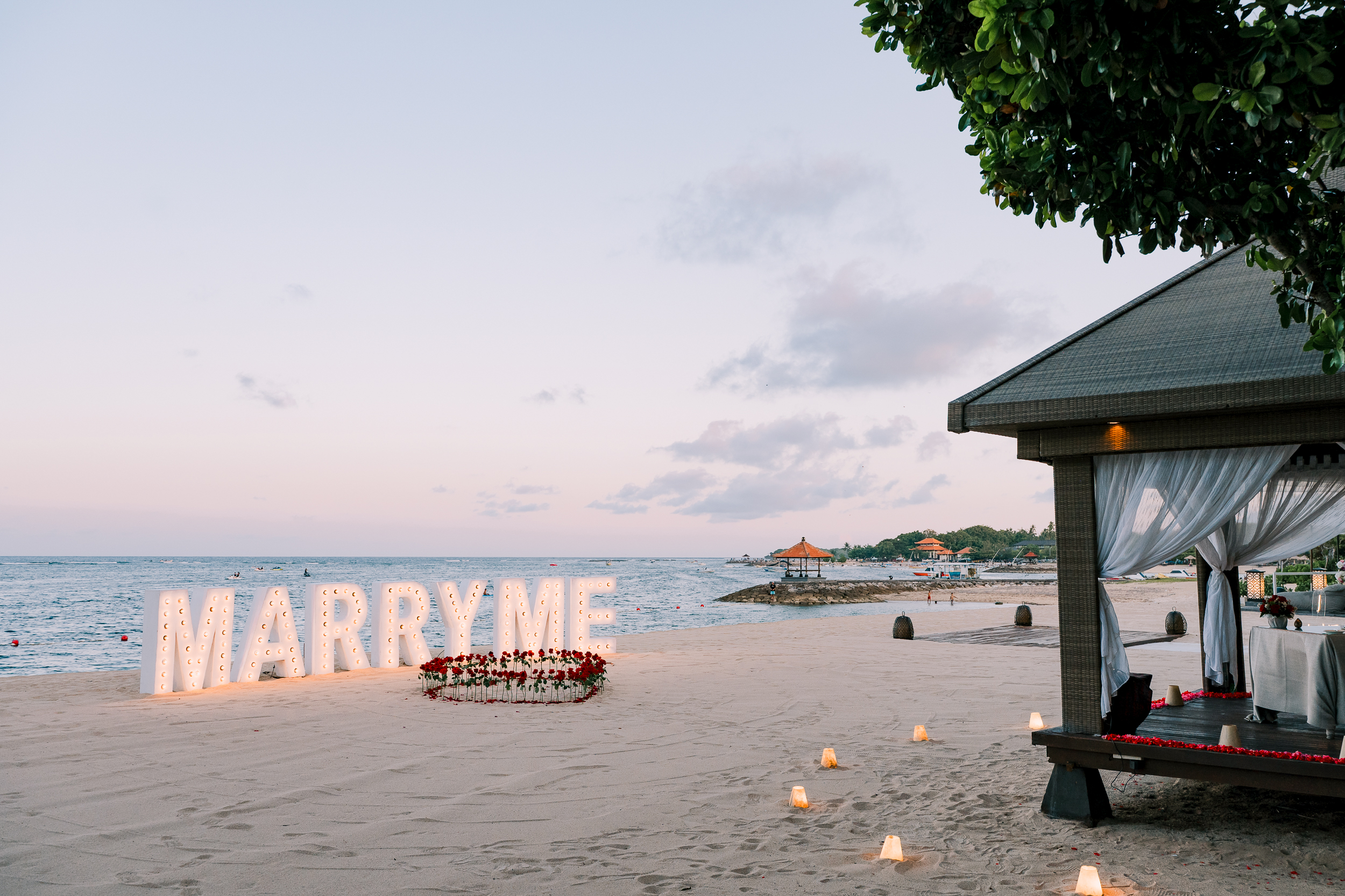 Illuminated Marry Me marquee letters and fresh red rose arrangement on a quiet oceanfront beach at dusk