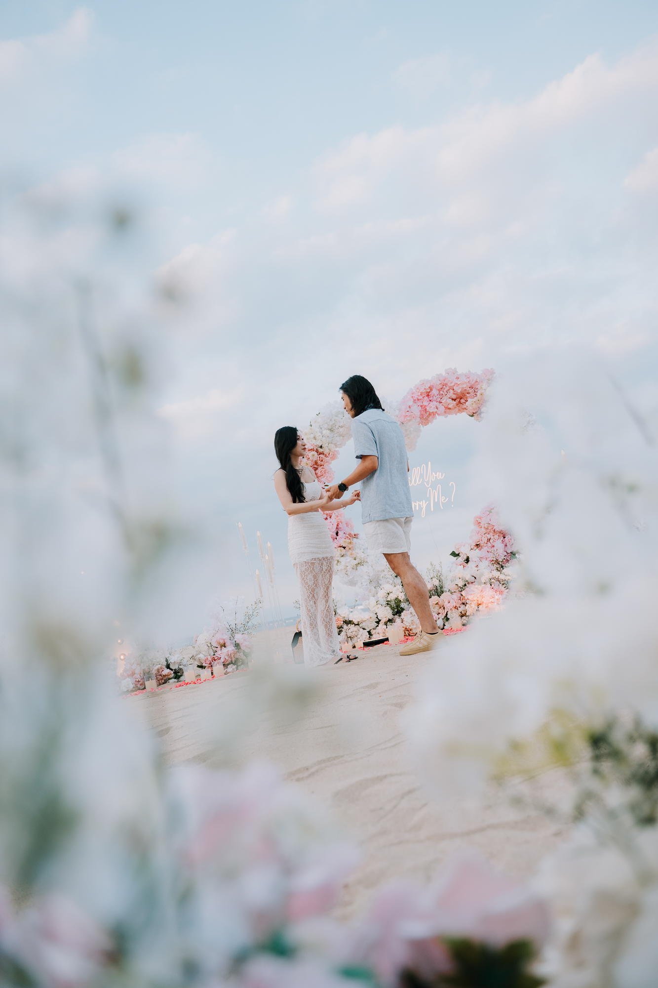 Framed shot through white flowers showing the couple standing together by the beach proposal setup