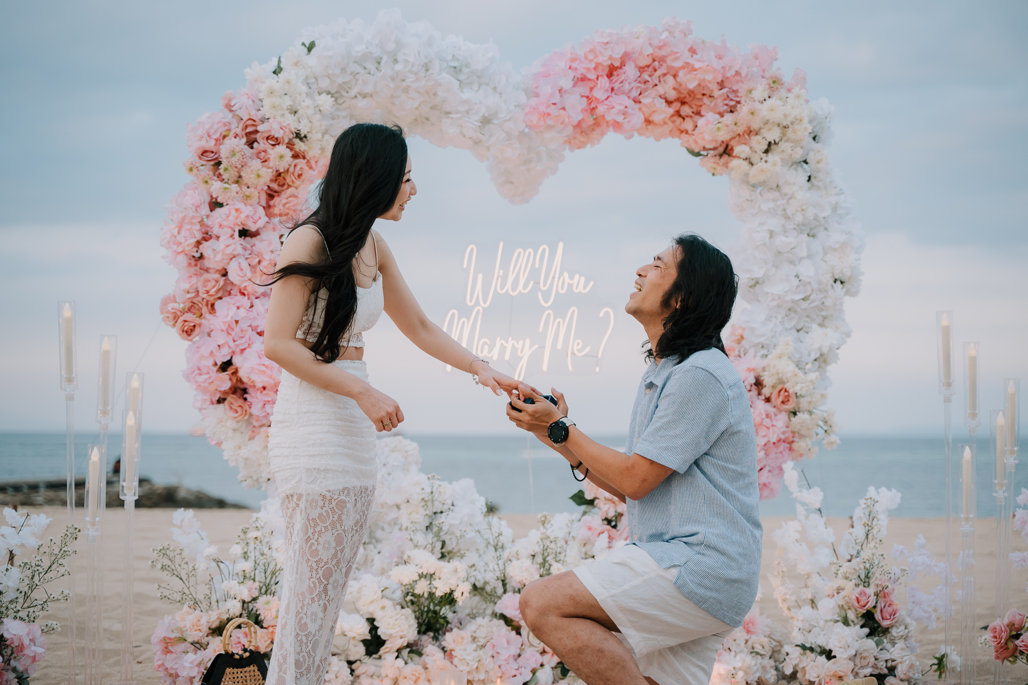 Smiling woman looking down at her partner kneeling with an open ring box on the sand
