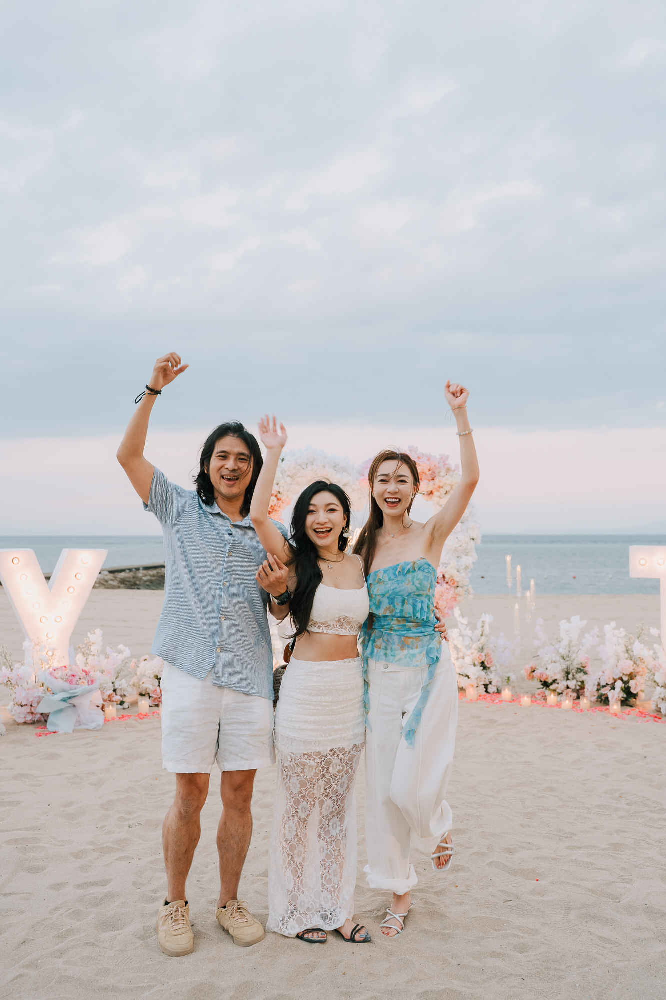Couple and friend raising their hands in celebration on the beach in front of glowing marquee letters