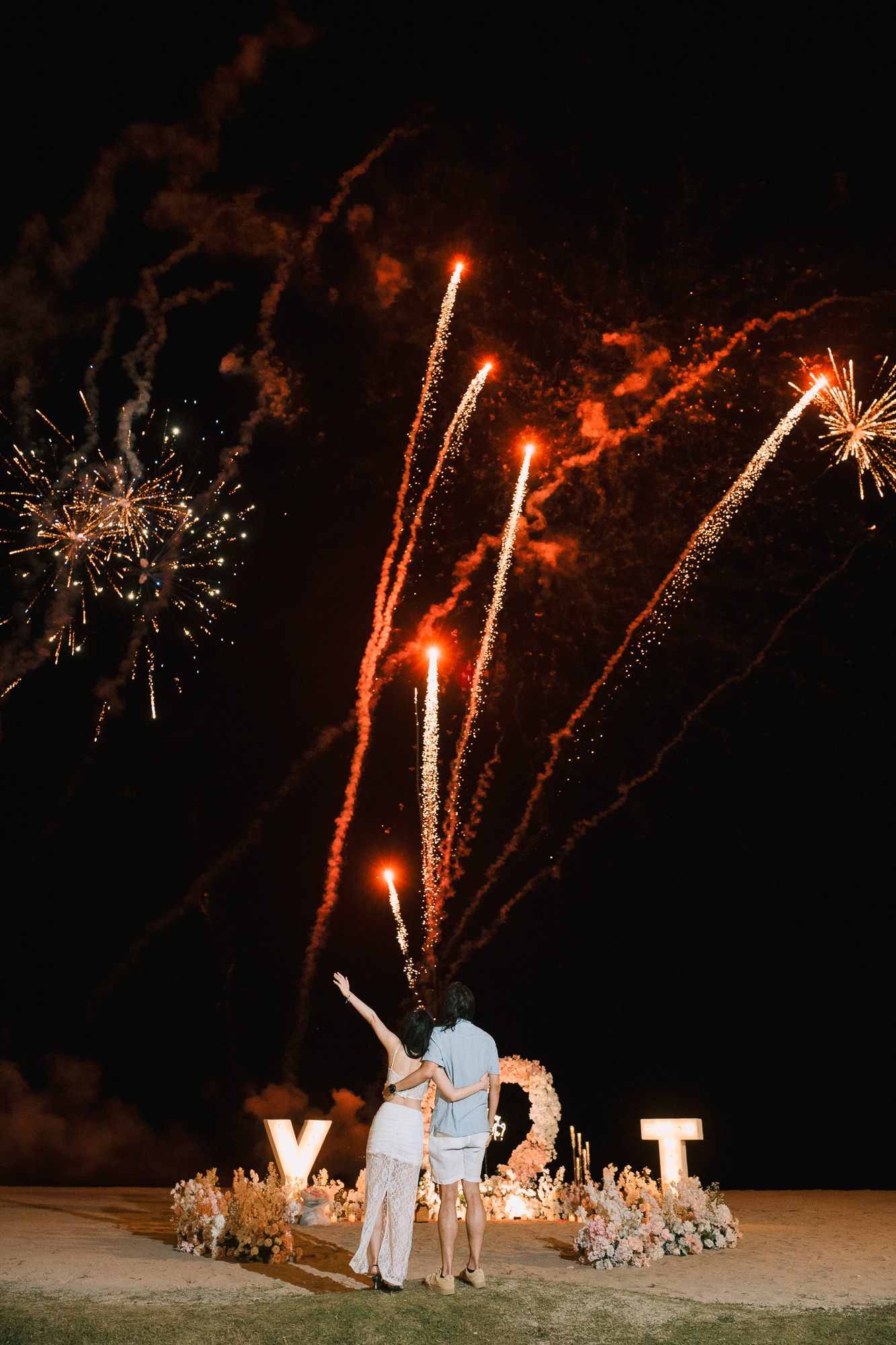 Rear view of a couple on a private Bali beach with woman raising her hand as bright red fireworks shoot into the night sky over glowing marquee letters