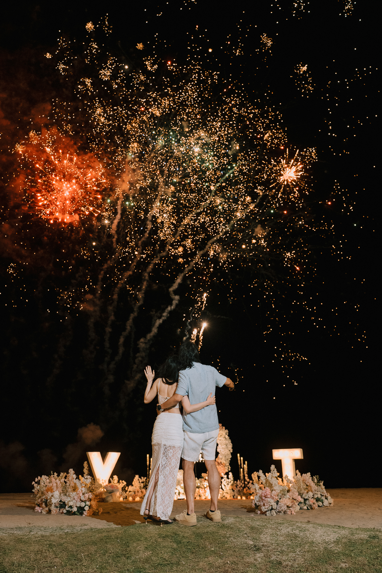 Newly engaged couple with arms around each other watching a golden firework explosion directly overhead on a dark beach