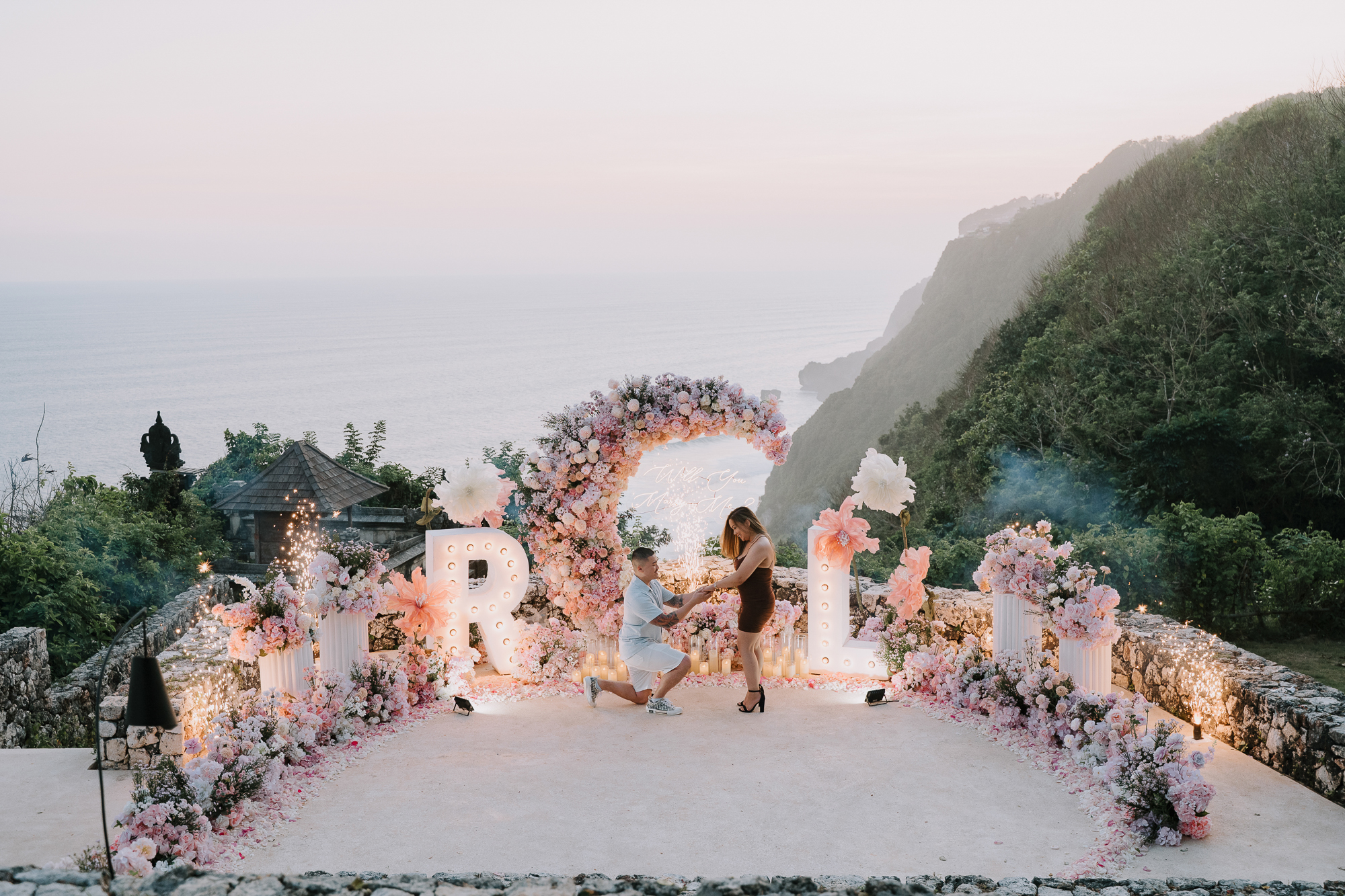 Groom dropping to one knee for a surprise helicopter proposal on a private Bali cliffside.