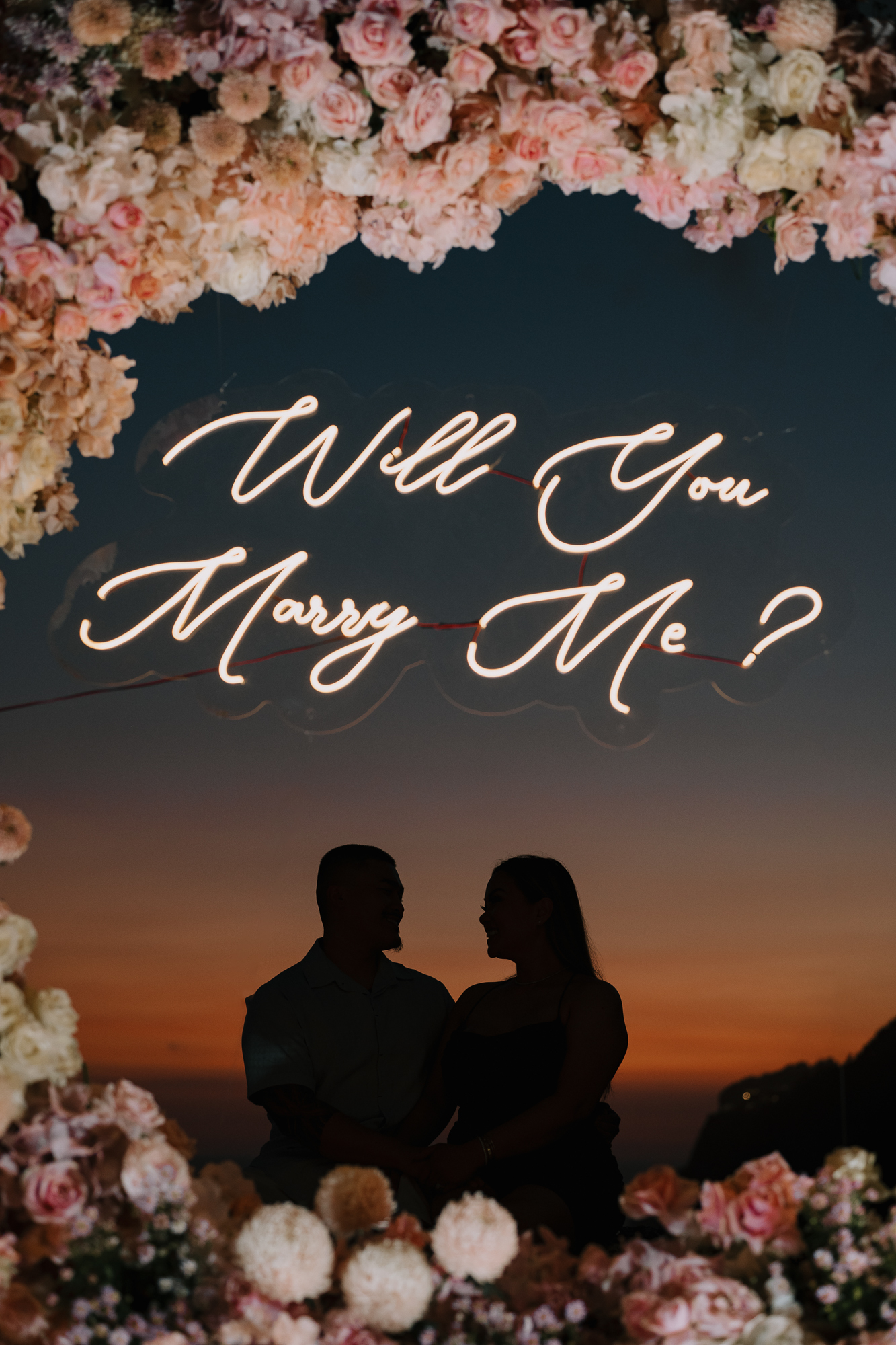 Dusk silhouette portrait of an engaged couple beneath a glowing Will You Marry Me neon sign