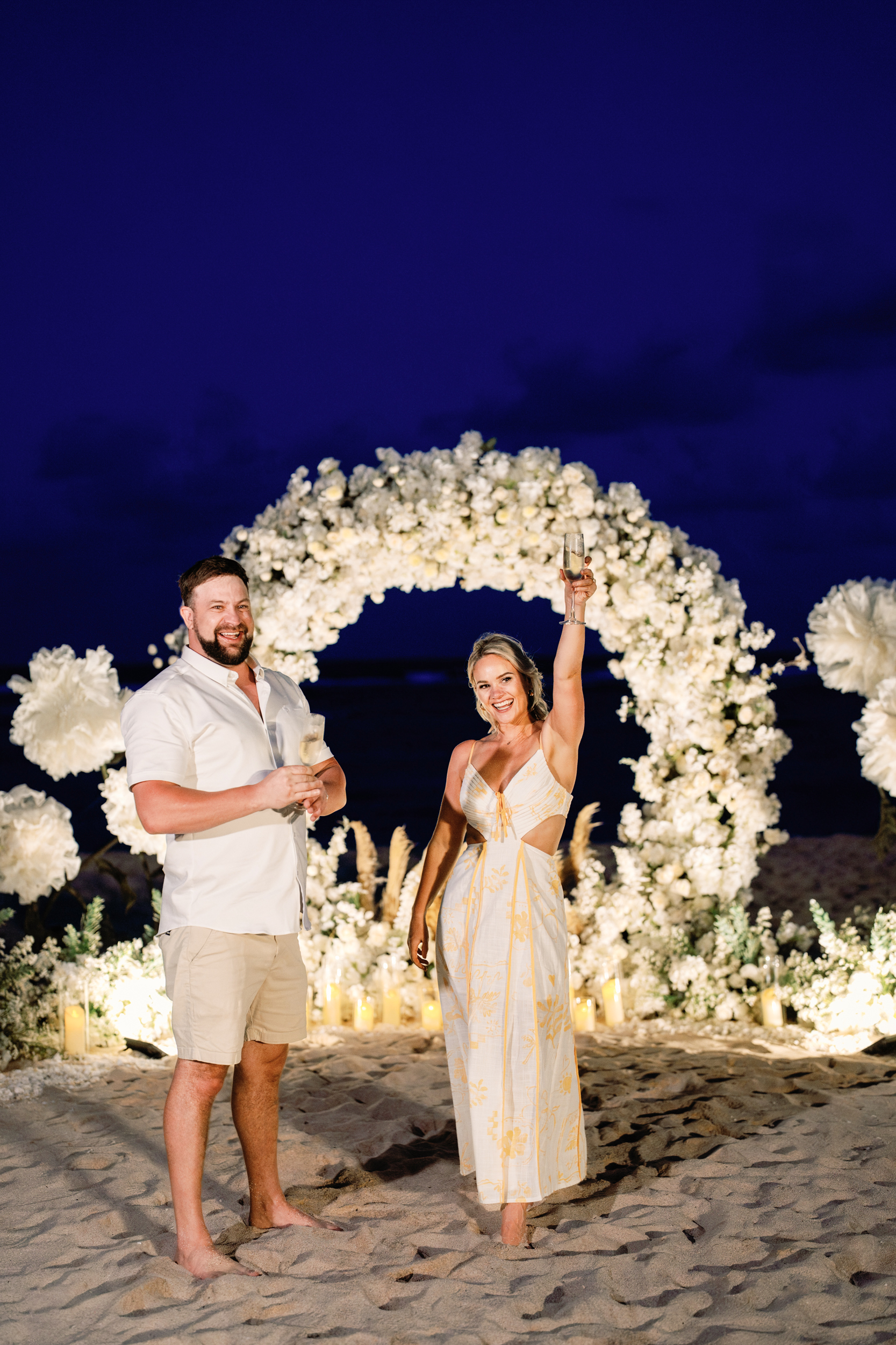 Night view of an illuminated white floral arch and candles on a private Bali beach proposal.
