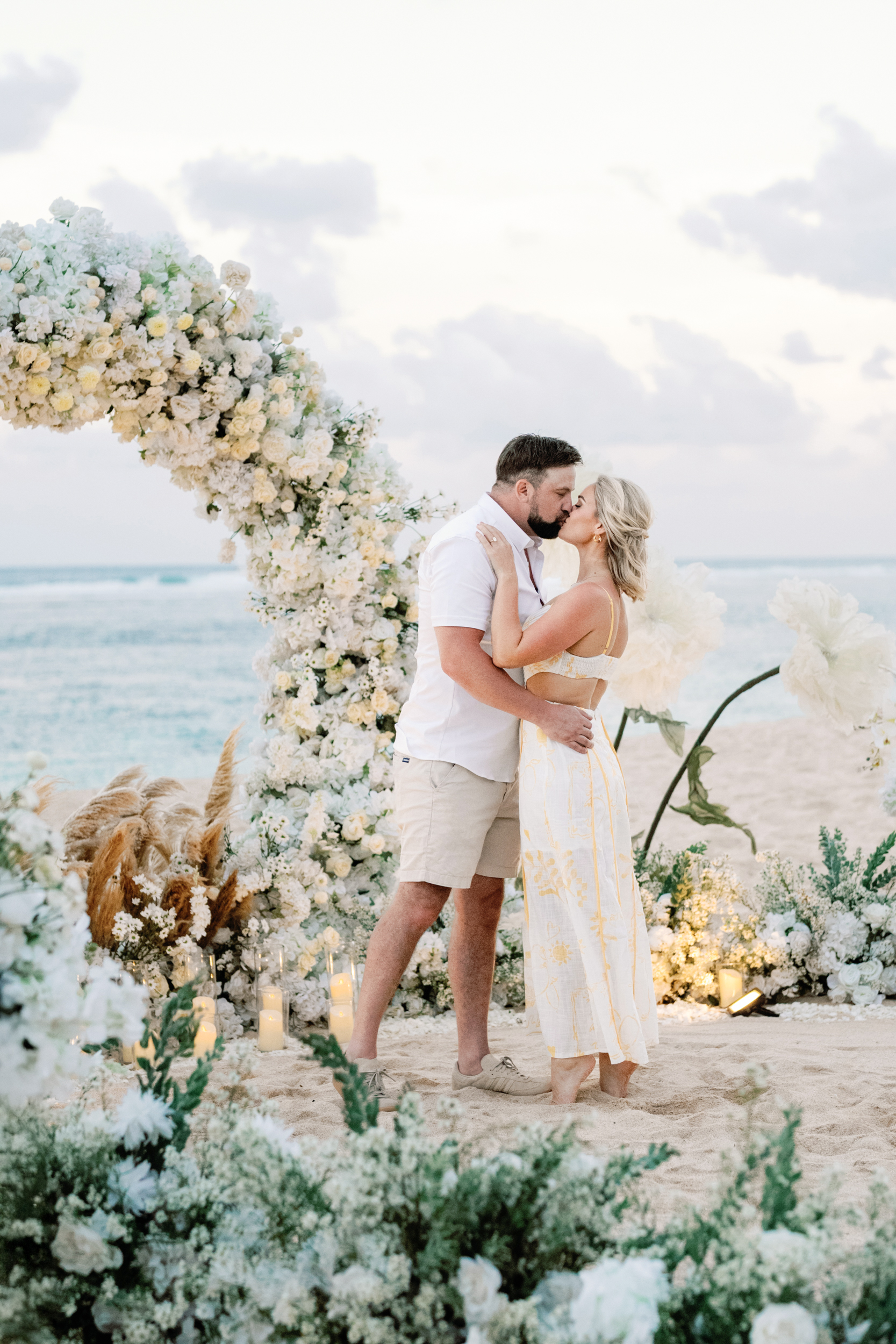 Newly engaged couple kissing in front of a white floral arch setup on a private Bali beach.