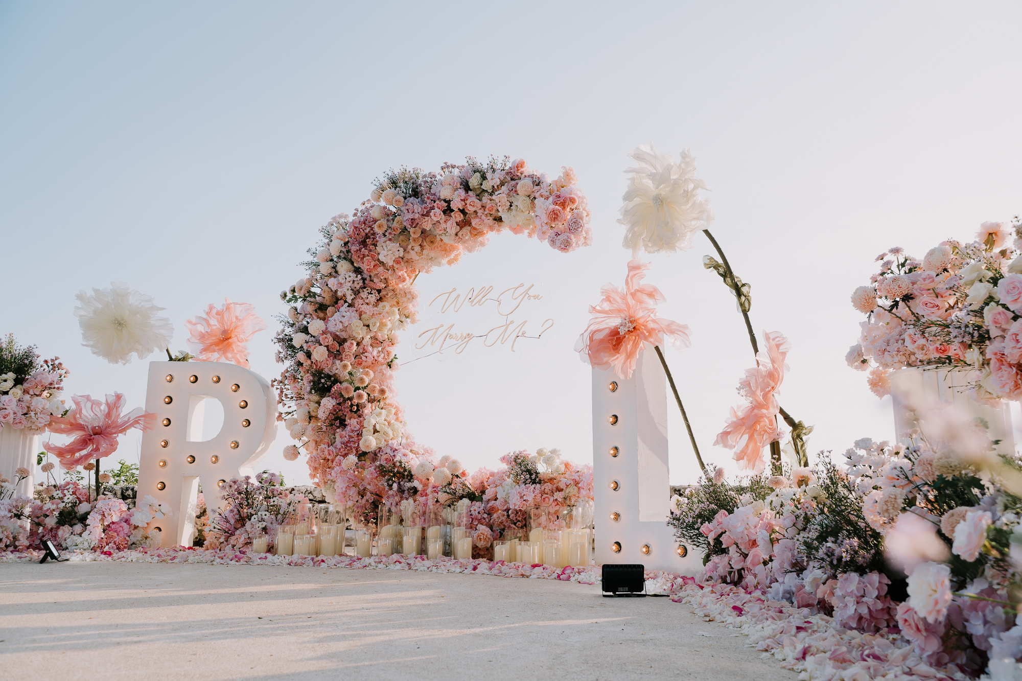 Panoramic view of a crescent floral arch installation, candles, and initial marquee letters on an Uluwatu cliff terrace