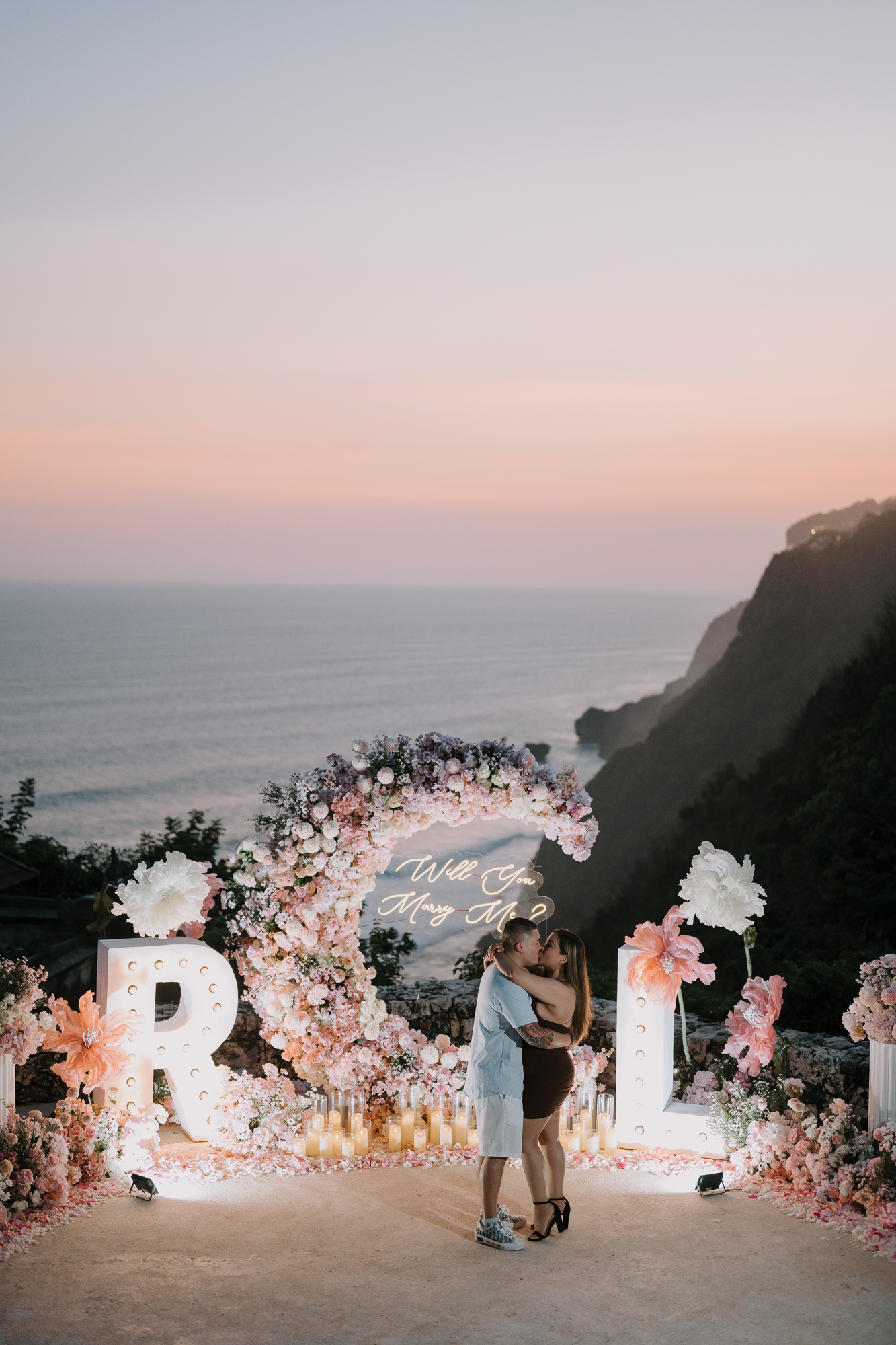 Sunset proposal moment framed by a blush crescent arch on an oceanfront Uluwatu cliff
