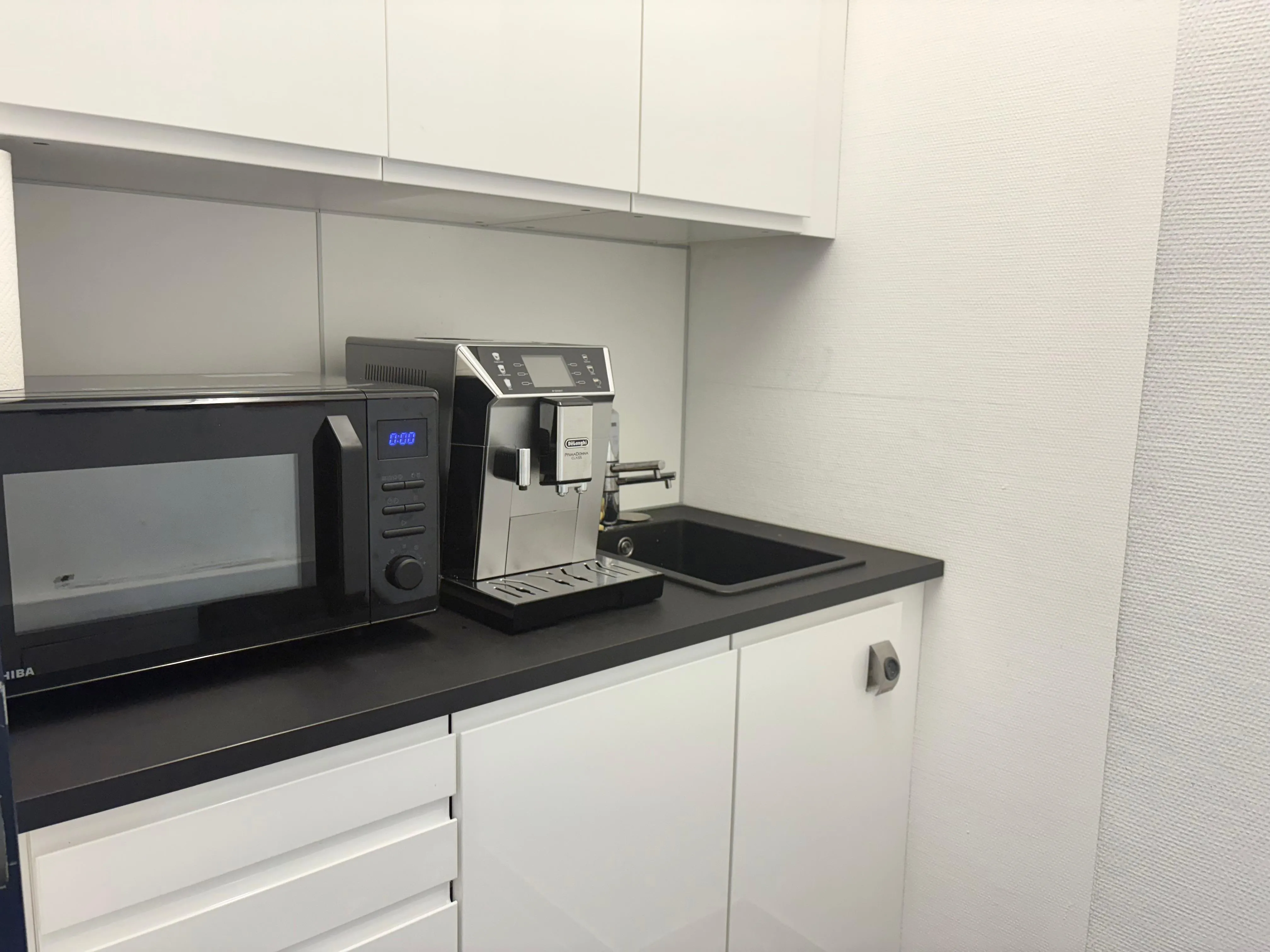 Moderne kleine Küchenzeile mit Mikrowelle, Kaffeevollautomat und Spüle auf schwarzer Arbeitsplatte.