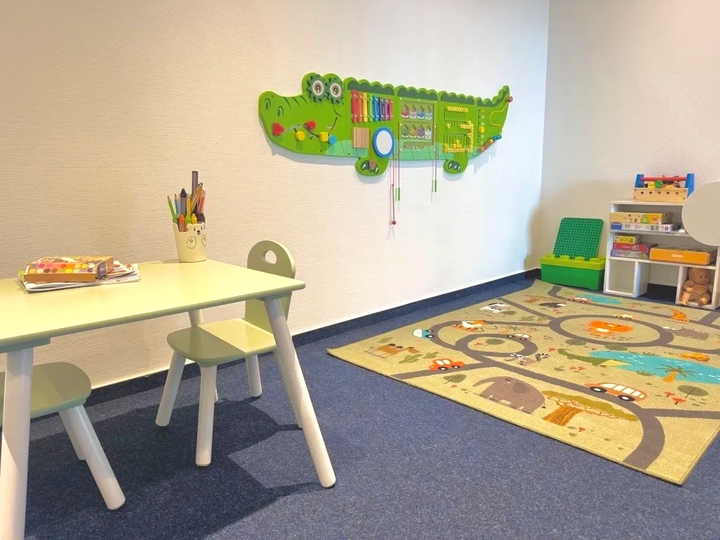Kinderspielzimmer mit zwei kleinen Stühlen und Tisch, buntem Teppich mit Tierspuren und grünem Krokodil-Wandspiel.