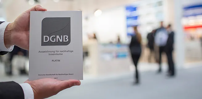 Zwei Hände halten eine Tafel mit DGNB-Platin-Auszeichnung für nachhaltige Innenräume vor unscharfem Hintergrund mit Menschen auf einer Messe.