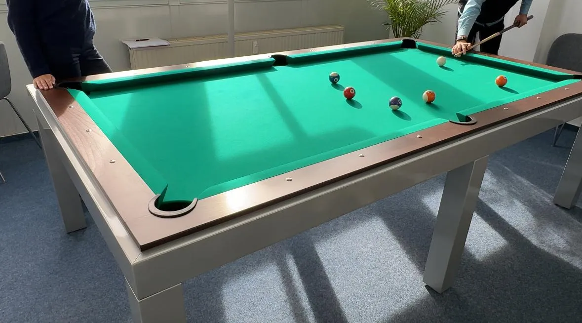 Zwei Personen, die an einem grünen Billardtisch mit mehreren Billardkugeln spielen, in einem hellen Raum mit blauer Teppichboden.
