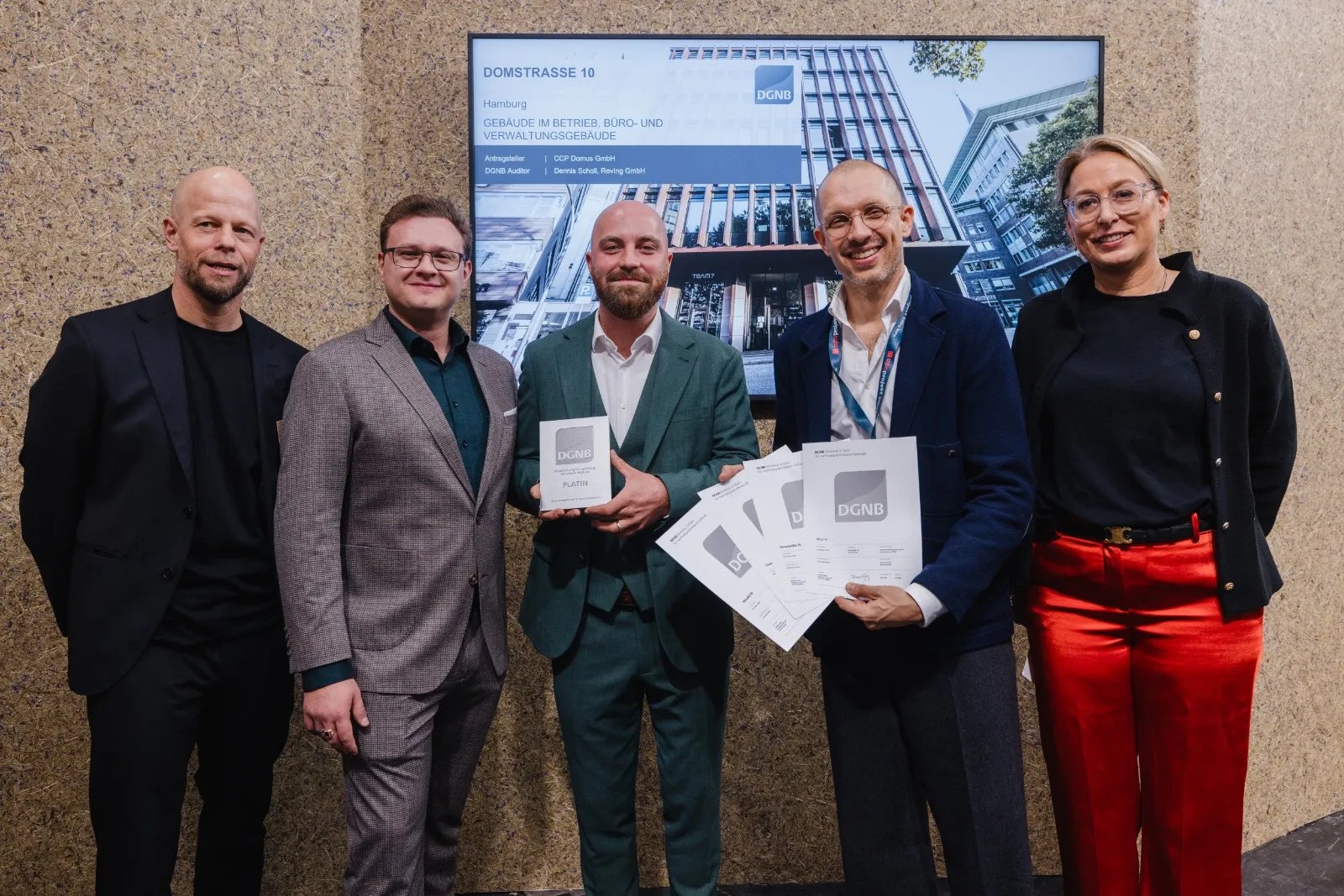 Fünf lächelnde Fachkräfte stehen gemeinsam in einem Innenraum, während zwei Männer in der Mitte offizielle DGNB-Zertifizierungsdokumente vor einem digitalen Bildschirm mit einem Immobilienprojekt hochhalten.