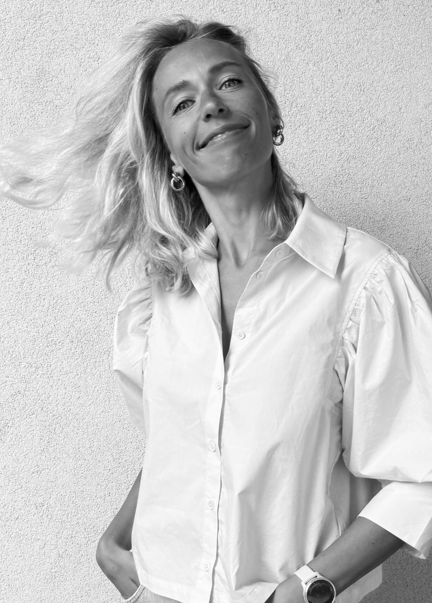 Smiling woman with wavy blonde hair wearing hoop earrings, a white button-up shirt, and a wristwatch, standing against a textured wall.