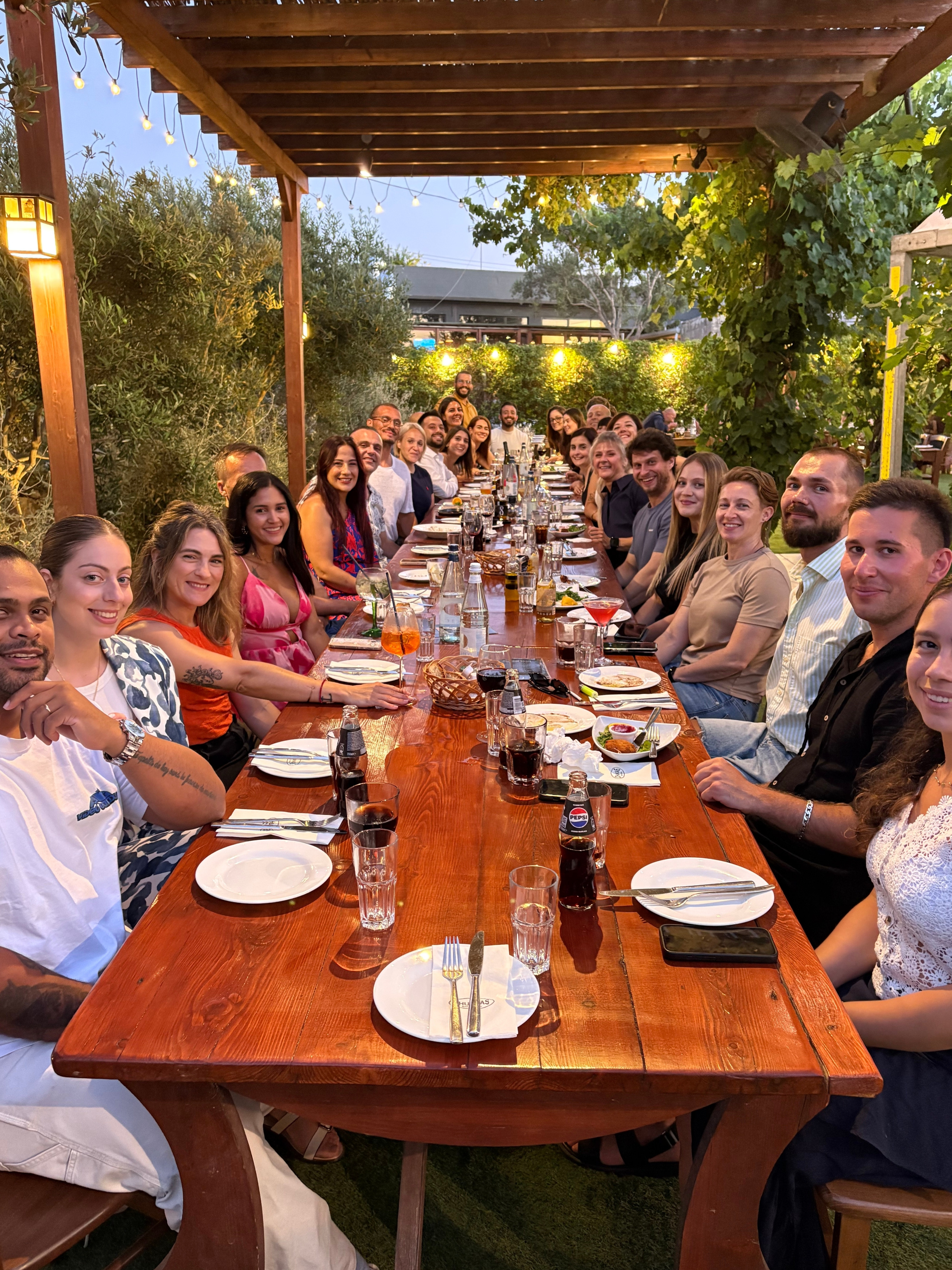 Members of CrossFit Martell gathered around a long outdoor dinner table in Malta, sharing food, drinks, and conversation during a community social event. The group smiles toward the camera under warm string lights and a wooden pergola, surrounded by greenery, highlighting the strong friendships and welcoming atmosphere that extend beyond the gym.