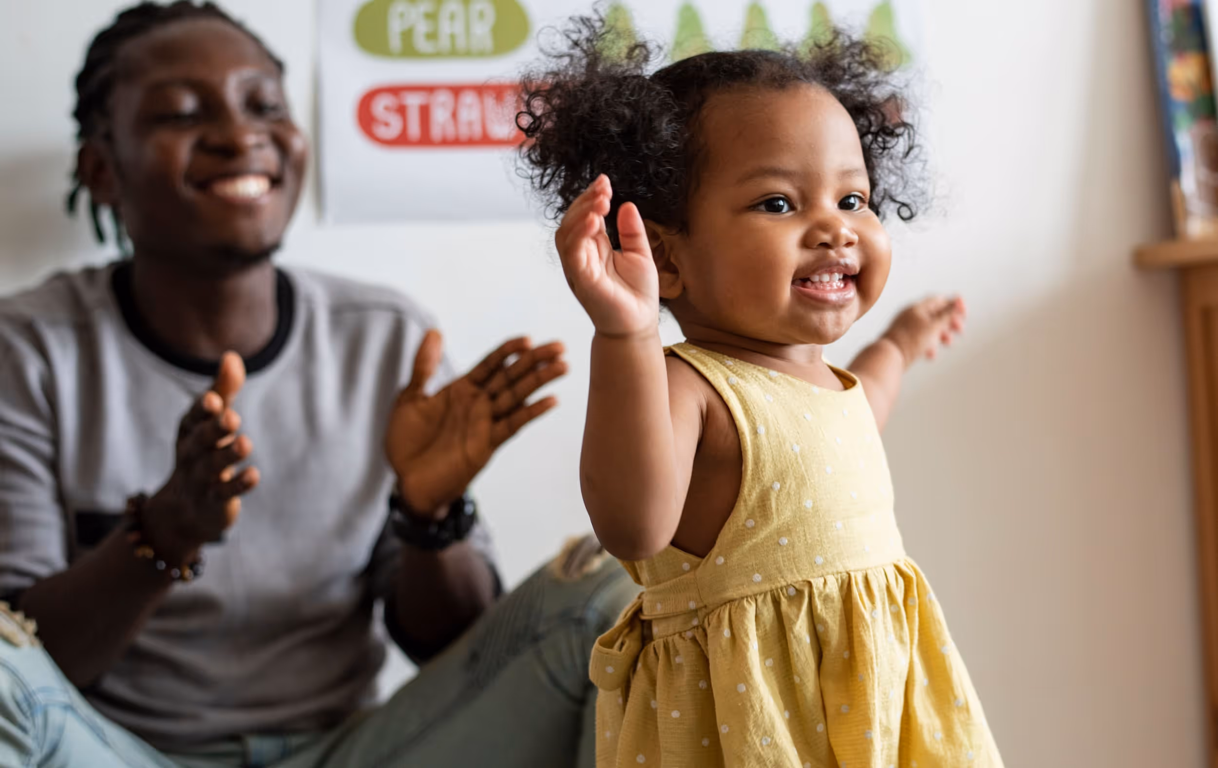 Happy black toddler with curly pigtails taking steps and dad celebrating her in the background. 