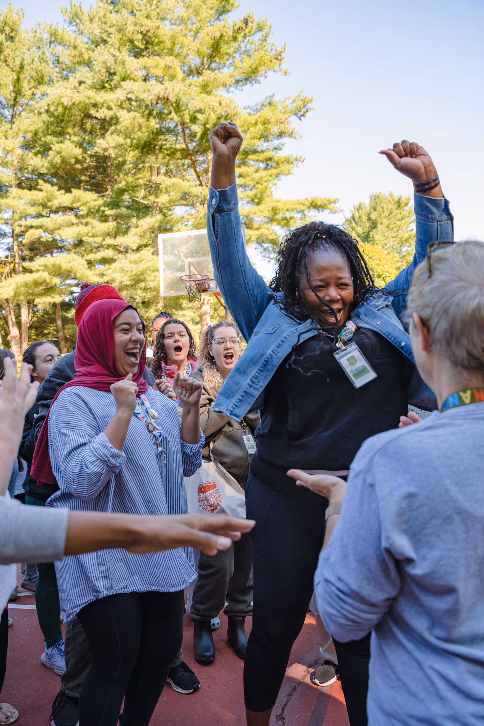 Diverse group of people celebrating together outdoors, with a woman in the center raising her arms in joy, surrounded by others with outstretched hands, on a basketball court with trees in the background.