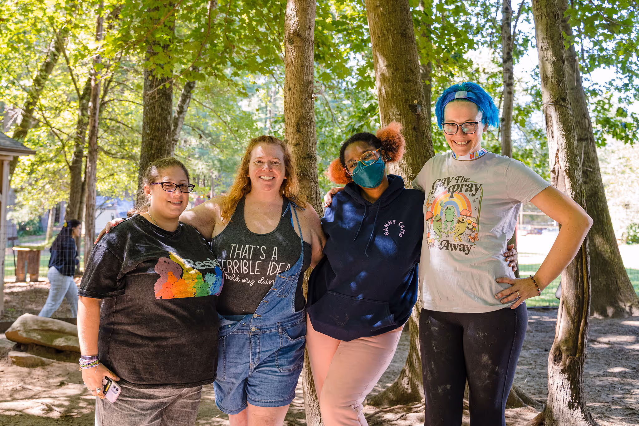 Four young women pose together in a wooded area, smiling and standing in casual outdoor attire, conveying a sense of friendship and fun, perfect for a nanny agency promoting positive relationships and activities for kids.