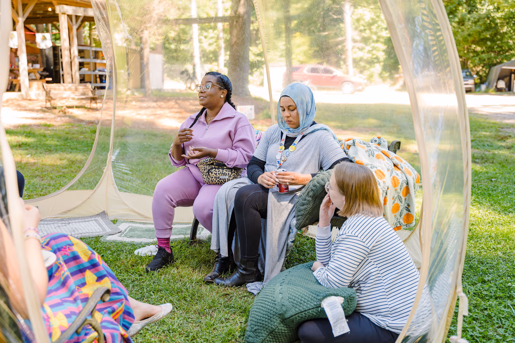 Diverse group of nannies sitting in a park, engaged in conversation, with a transparent dome structure around them, conveying a sense of community and connection among childcare professionals in a natural setting.