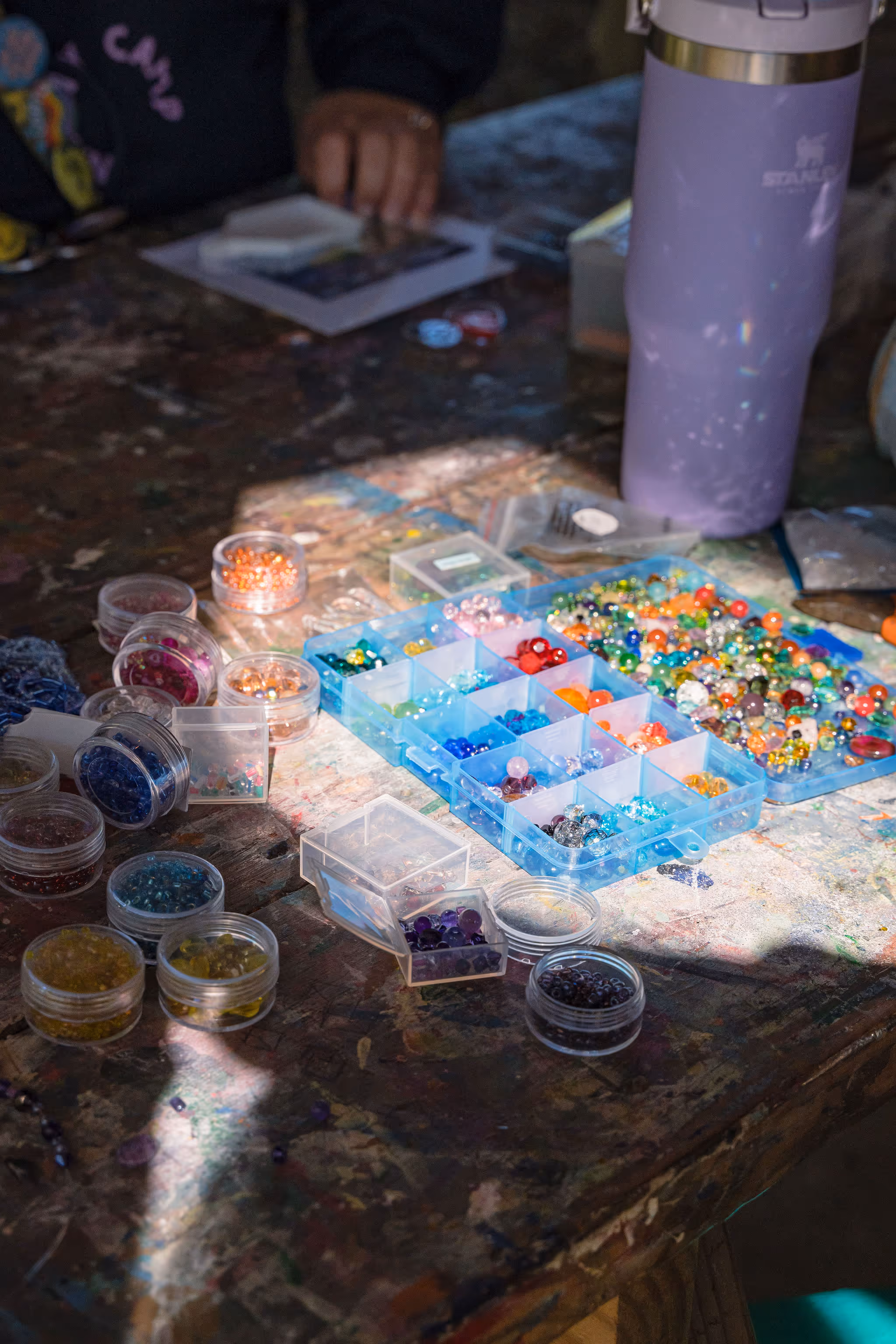 Beads and containers on a table with a child, showcasing a creative activity. Colorful beads in various containers, a purple water bottle, and a child's hands visible in the background, engaged in an arts and crafts project.