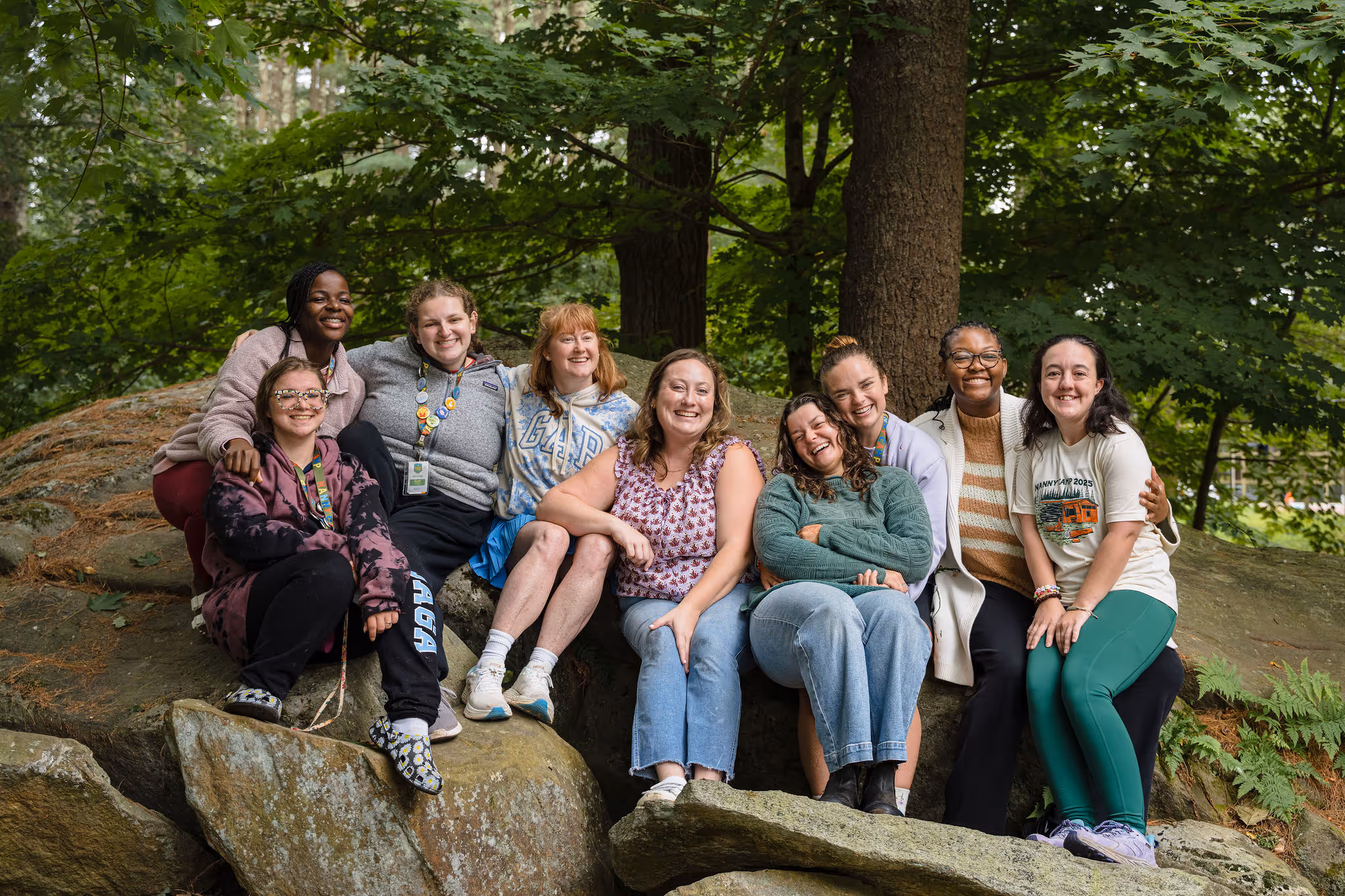 Nine women sitting together on large rocks in a forest, smiling and posing for a group photo, showcasing diverse group of nannies or caregivers enjoying outdoor activities.