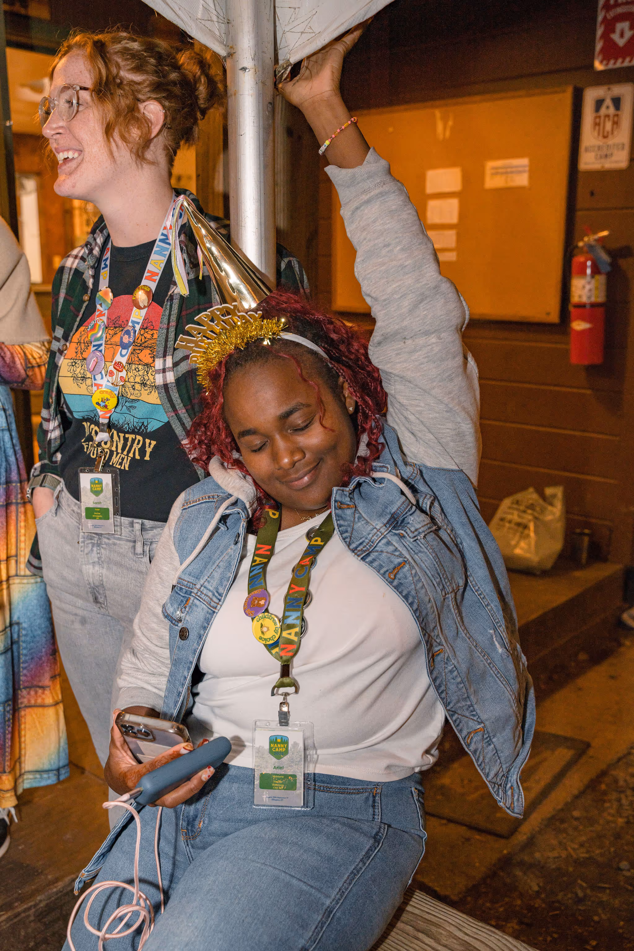 Two young women, one sitting and one standing, wearing casual clothes and party accessories, smiling and having fun together, with a fire extinguisher and bulletin board visible in the background.