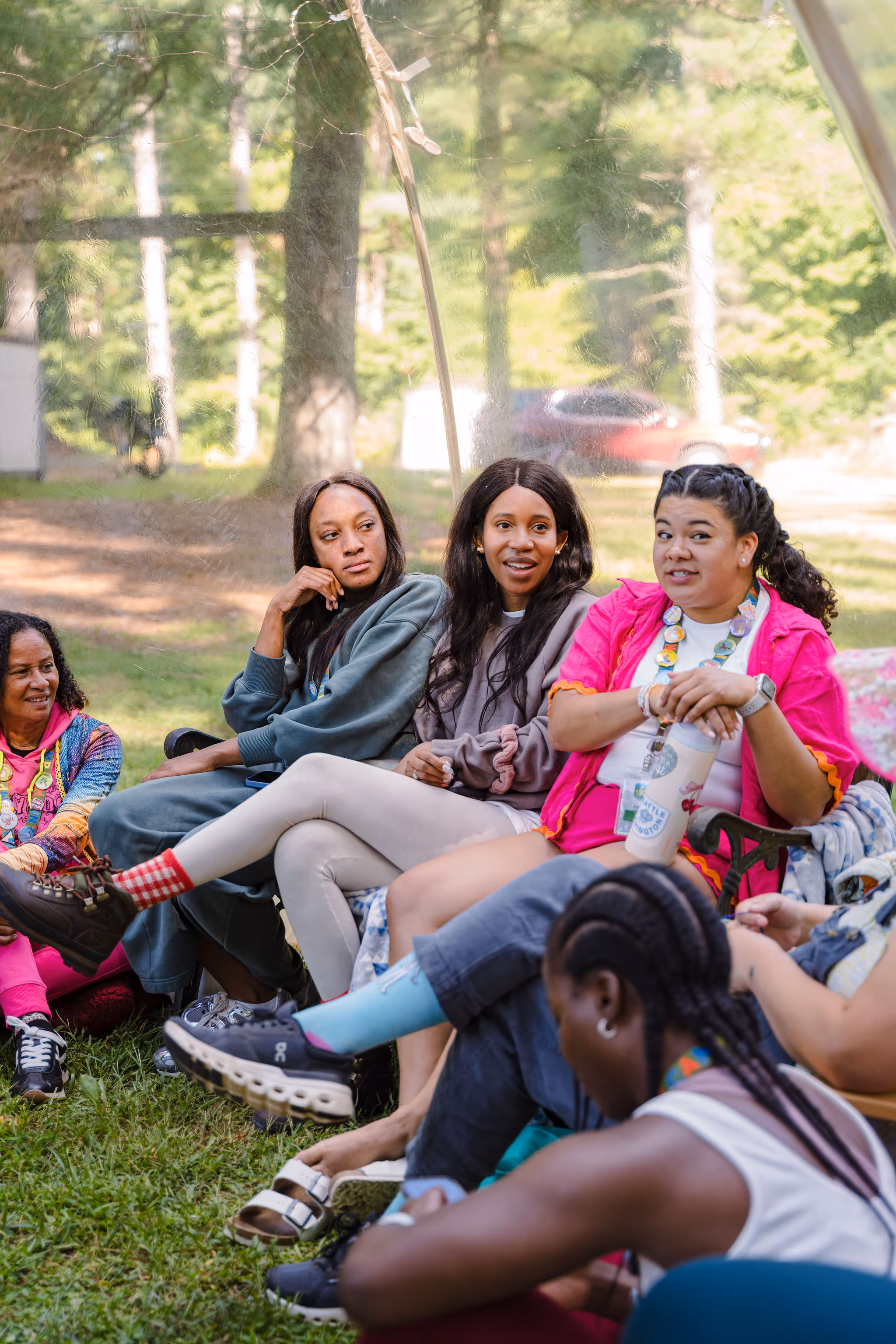 Diverse group of nannies gathered outdoors, sitting on grass in a relaxed setting, engaging in conversation and enjoying each other's company in a natural, serene environment.