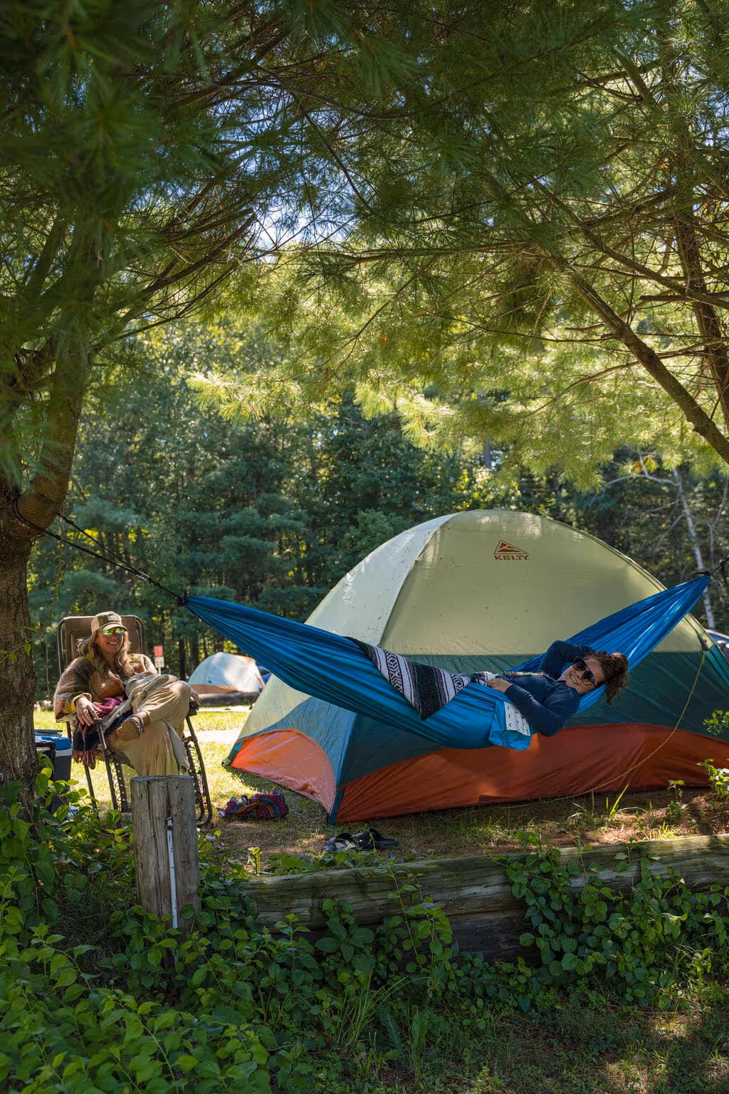 Woman relaxing in a hammock with another person nearby, sitting in a chair, amidst camping gear and a tent in a wooded area, surrounded by trees and greenery, enjoying the outdoors together.
