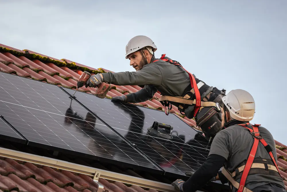 Foto von Solarmonteuren auf dem Dach. Ein Monteur lehnt sich über das Modul, um die Modulklemme zwischen zwei Modulen mit einem Akkuschrauber festzuziehen.