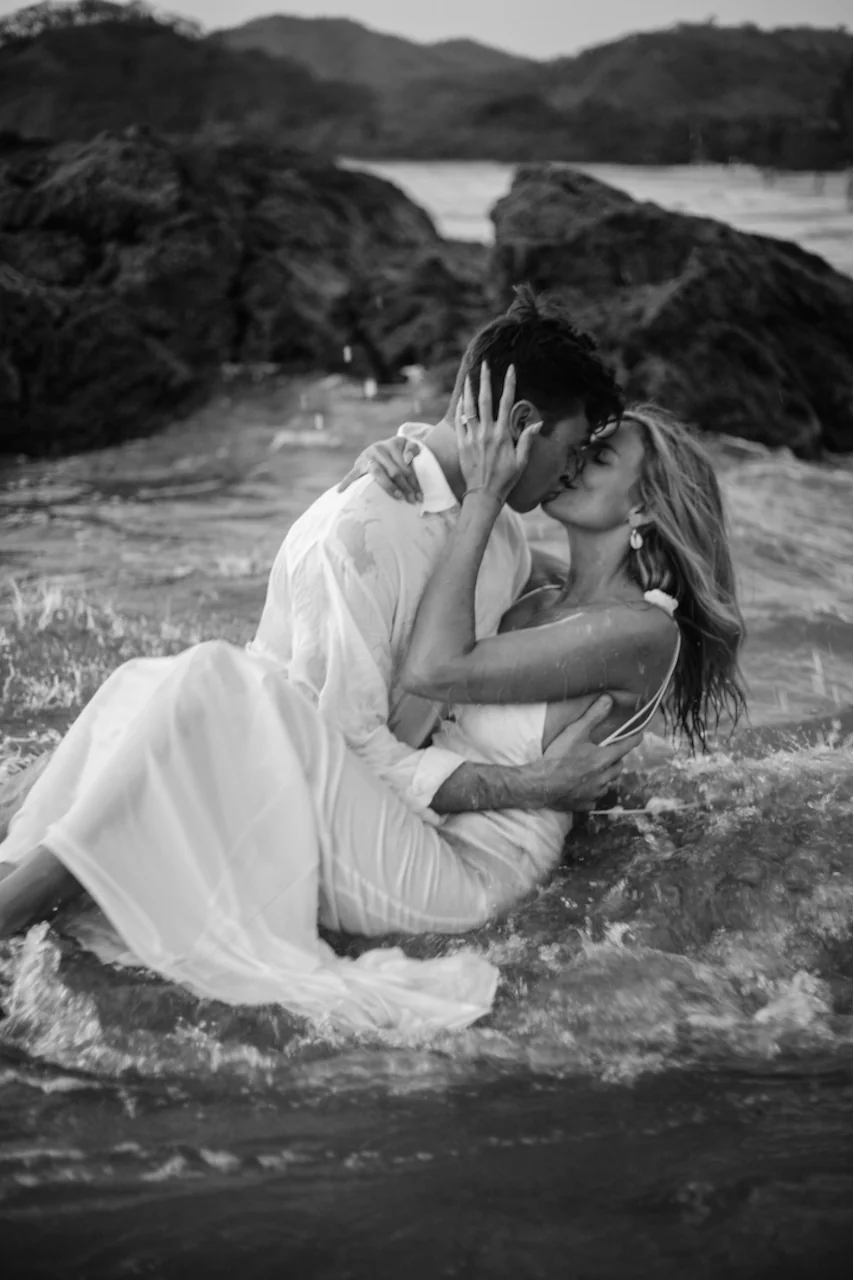 Couple in white embracing in the ocean shallows during a black-and-white portrait session on a Costa Rica beach