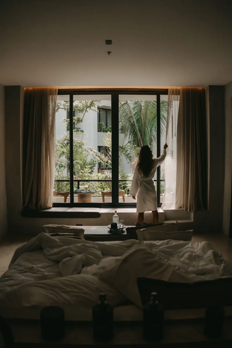 Woman in a robe opening curtains to a tropical morning, unmade bed in the foreground