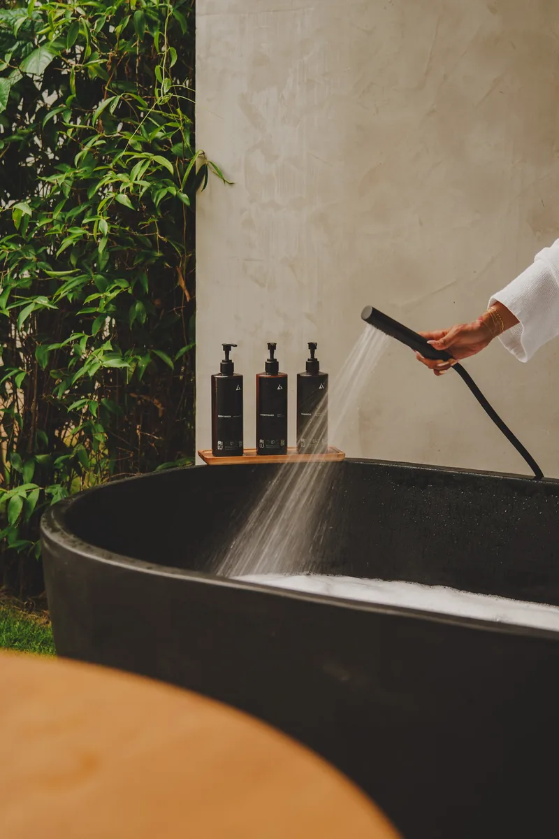 Branded bath products on a wood tray beside a stone soaking tub in a tropical Costa Rica setting