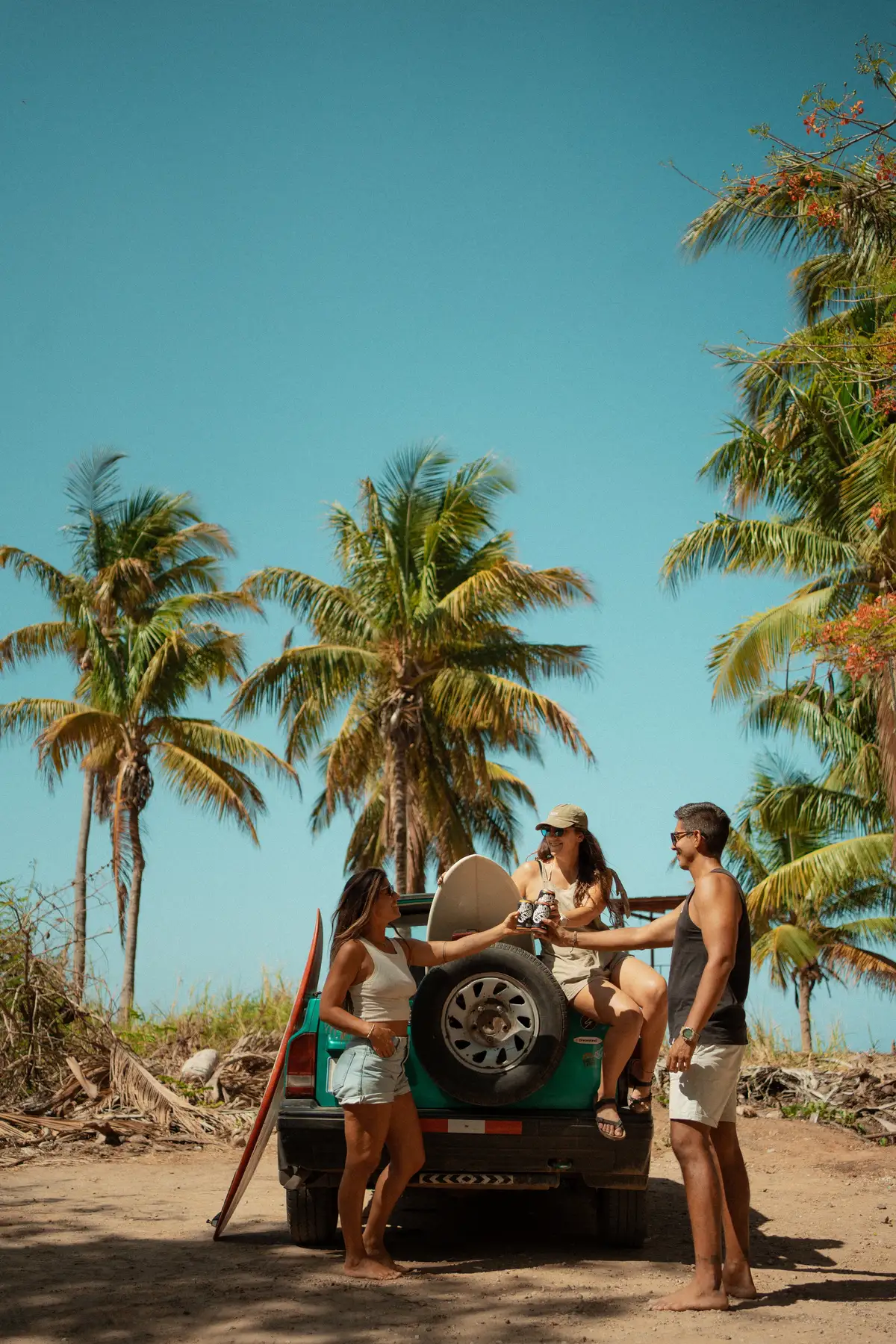 Three friends toasting drinks by a surf truck under palm trees on a Costa Rica beach, authentic user-generated content moment