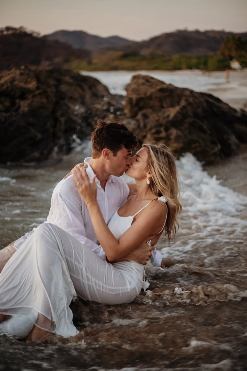 Couple in white kissing in the surf between dark volcanic rocks at sunset during beach engagement photos in Guanacaste, Costa Rica