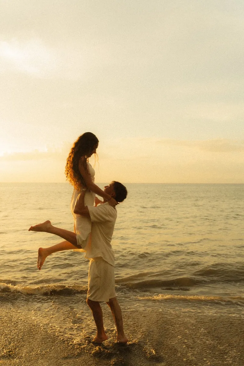 Man lifting his partner at the water's edge at golden hour, both in flowing cream linen, an example of what to wear for beach engagement photos in Costa Rica